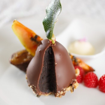 Photo of a dessert display resembling a chocolate pear shaped cake with a leaf at the top garnished with raspberries and a slice of papaya 