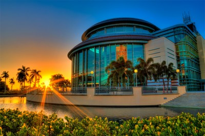 Photo of Large round glass building with stairs and a concrete pond located in front. A bright orange sunset and clear blue sky left of the building.