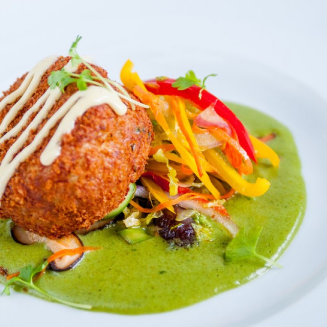 Photo of a Fried crab cake leaning on a julienne mix of red, orange and yellow peppers, on a green sauce