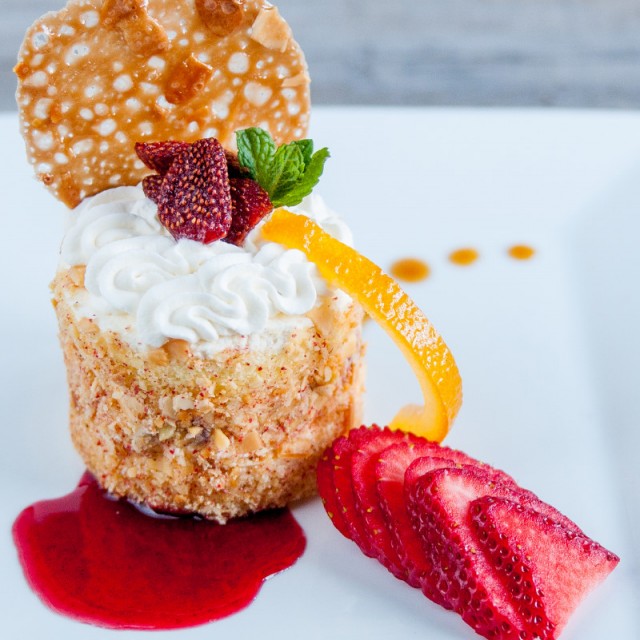 Food Photo of round piece of cheesecake with nut and graham cracker crusting on a bed of raspberry sauce, whipped cream swirl, candied orange peel, and sliced strawberries.